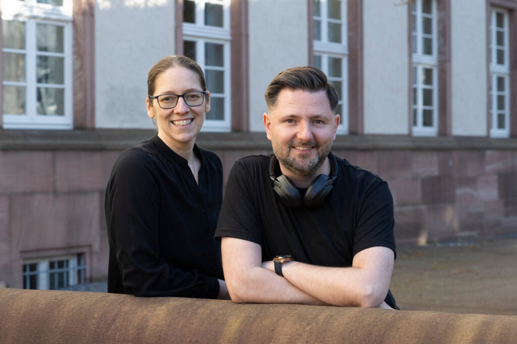 Zwei Mitglieder des Podcast-Teams des Hessischen Landesarchivs, Jan-Hendrik Evers und Maria Kobold, stehen vor einem historischen Archivgebäude und blicken lächelnd in die Kamera.